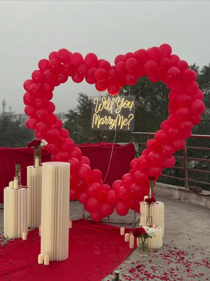 I love creating bold and romantic statements, like this stunning terrace proposal setup. A giant heart-shaped arch made of red balloons creates a dramatic frame for the 'Will You Marry Me?' neon sign, while the red carpet and candles make the moment feel like a movie scene.