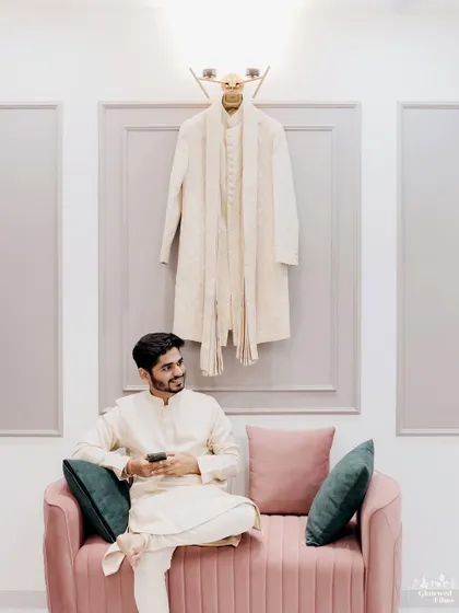 The groom, Mahek, relaxes on a pink sofa, looking at his sherwani hanging on the wall, a quiet moment before the festivities.