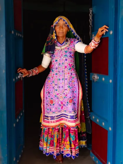 A Meghwal woman in a stunningly embroidered purple dress stands in the doorway of her home. The vibrant colors of her attire and the blue door create a beautiful composition.