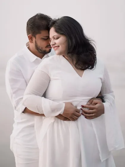 An intimate hug from behind on the beach. The all-white outfits and the soft, overcast light create a very romantic and serene mood.