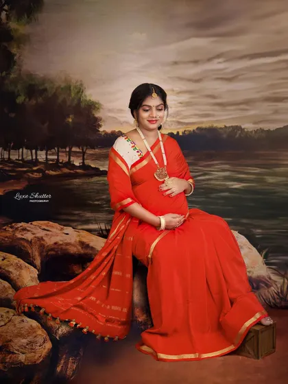 A beautiful, painterly portrait of a mother-to-be in a red saree, seated by a riverbank. The style is reminiscent of classic Indian art, creating a timeless and soulful image.