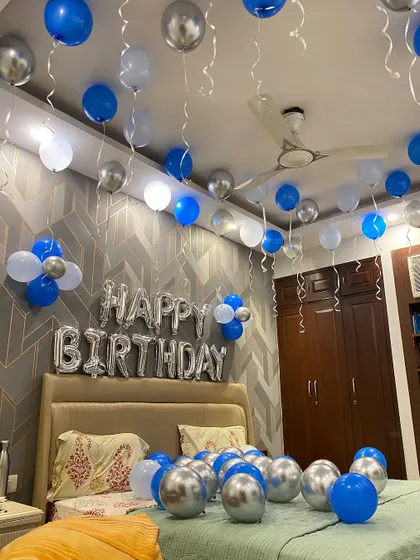 A simple yet stylish birthday room decoration. This setup includes blue, white, and silver chrome balloons on the ceiling and floor, with large "Happy Birthday" foil letters above the bed.