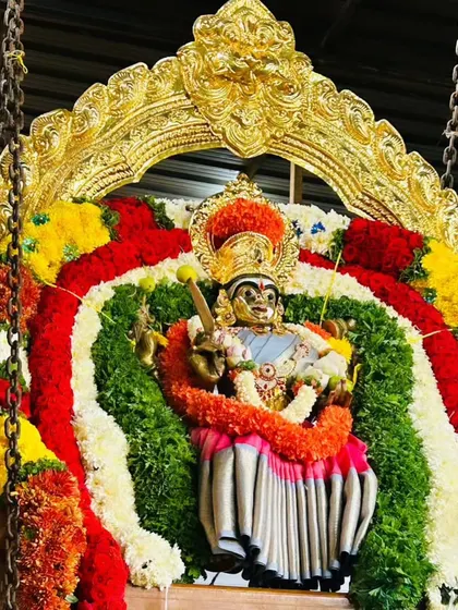 A deity on a swing, framed by a golden arch and decorated with thick, vibrant garlands of red, white, and yellow flowers.