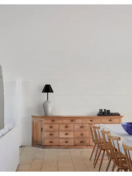 A table lamp from the 'Bernard and friends' collection sits atop a rustic wooden credenza. The lamp's simple, elegant form and natural wood texture bring a sense of calm and craftsmanship to the dining space.