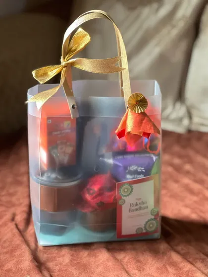 A festive Rakhi gift bag filled with snacks and a drink, finished with a bright orange decorative element and a gold ribbon.