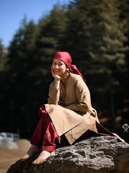 A full-length portrait of the woman sitting on a boulder, showcasing her complete traditional outfit against the backdrop of a sunlit forest.