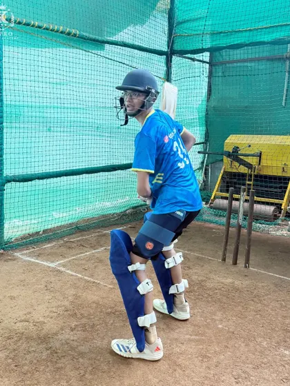 A young batsman practices his stance in the outdoor nets, with the bowling machine visible in the background, ready for a high-intensity session.