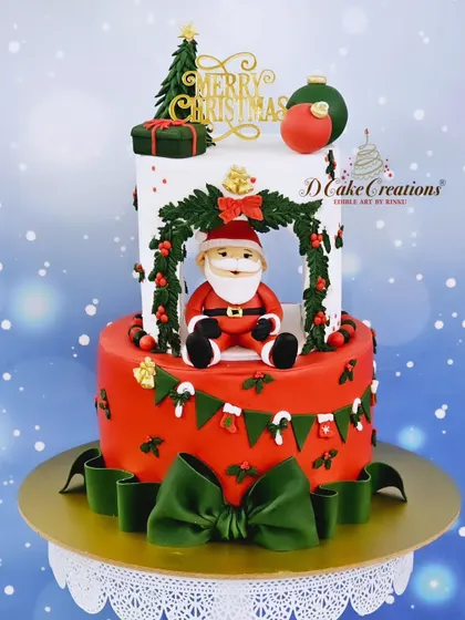 A fun and festive Christmas cake featuring a Santa figurine sitting inside a cutout tier. The cake is decorated with edible presents, a Christmas wreath, and a large green bow.