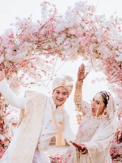A fun, candid moment of the couple striking a pose under a beautiful cherry blossom arch. Their coordinated white outfits and joyful expressions perfectly capture the lighthearted and celebratory spirit of their wedding day.