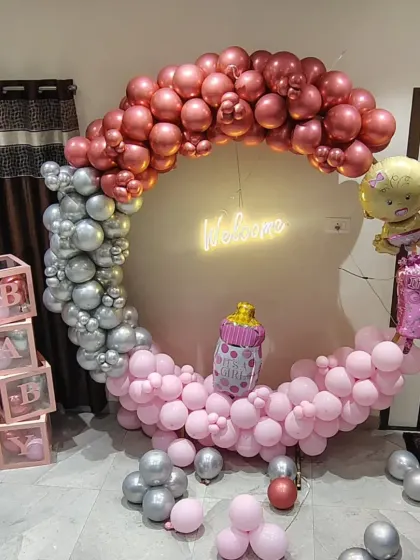 A "Welcome" baby girl decoration with a circular balloon arch in rose gold, pink, and silver, and "BABY" blocks.