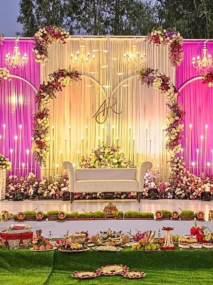 A fairytale engagement setup with a heart-shaped floral arch and monogram. The stage is bathed in warm light from hanging chandeliers and draped in pink and white fabrics, creating a romantic focal point.