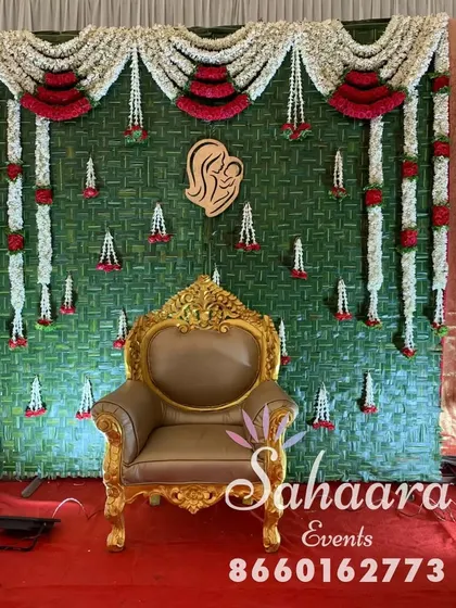 A close-up of our traditional baby shower backdrop. The design features a woven green mat, garlands of jasmine and roses, and a wooden cutout symbolizing motherhood.