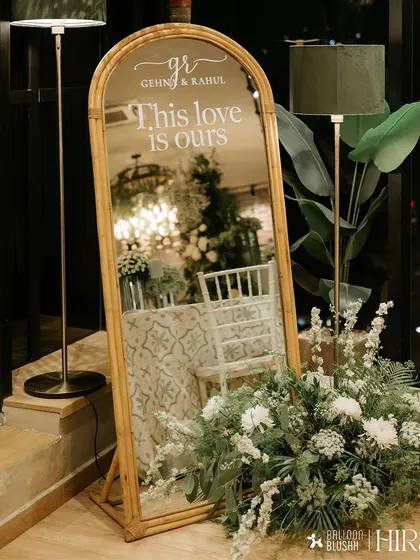 We love adding personal, reflective touches to our decor. This elegant arched mirror features a custom decal with the couple's names and the phrase "This love is ours," surrounded by a delicate arrangement of white flowers and greenery.