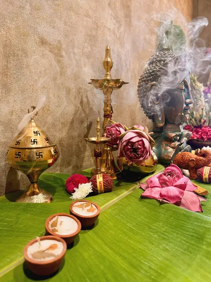 A sacred corner featuring traditional puja items. A brass incense burner sends fragrant smoke over a Buddha statue, lotus flowers, and diyas set on a fresh banana leaf.