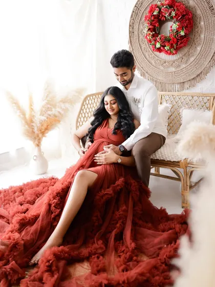A tender moment in a beautiful boho setting. The dad-to-be embraces his partner from behind, both of them looking down at the baby bump with love.