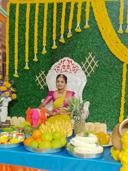 A vibrant half-saree function setup. The backdrop is a green wall decorated with hanging yellow flower garlands, paired with a comfortable throne for the girl of the hour.