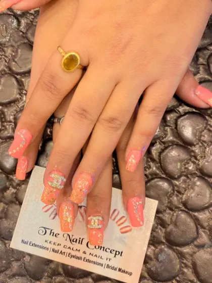 A fun and trendy set of peach-colored square nails. The design features glitter, crystal accents, and 3D Chanel logo embellishments.