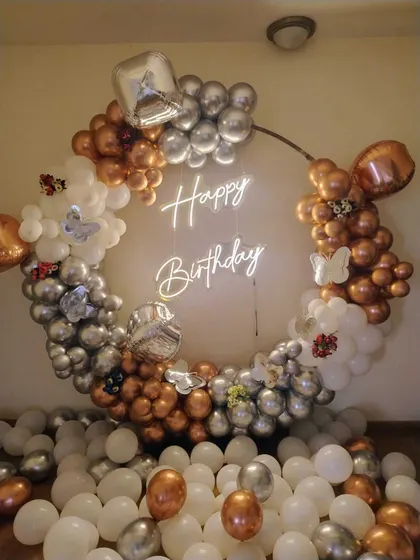 A birthday balloon ring with copper, silver, and white balloons. The setup includes butterfly accents and a neon sign, with loose balloons covering the floor.