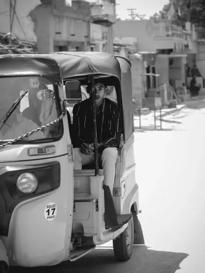 A candid shot of a young man in an auto-rickshaw, looking directly at the camera. The black and white edit gives it a timeless, classic feel.