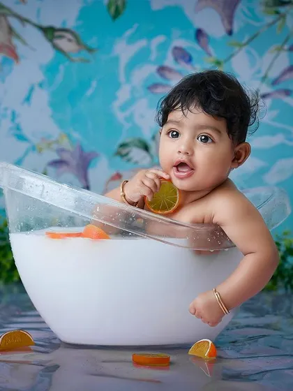 A fun and unique toddler "milk bath" session. The baby sits in a small tub with orange slices against a vibrant blue floral background, creating a playful and artistic image.