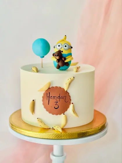 A fun Minion cake for a third birthday. This design features a Minion holding a teddy bear, surrounded by his favourite snack, bananas, on a simple buttercream cake with a personalized name plaque.