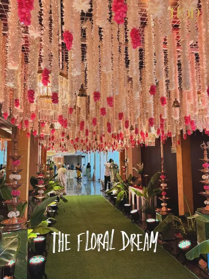 A wide shot of "The Floral Dream" entrance aisle. The green carpet provides a beautiful contrast to the pink and white floral ceiling, and the path is lined with traditional brass lamps for a warm glow.