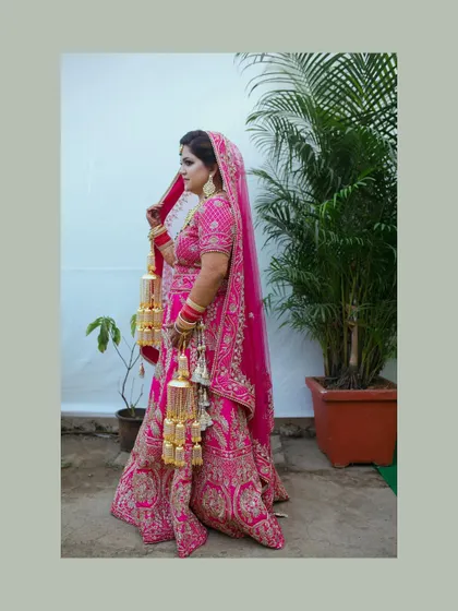 A full-length portrait of a bride in a vibrant pink lehenga. The shot showcases her entire outfit, including the beautiful kalire, against a simple background.