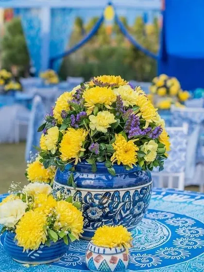 A close-up of a table centerpiece, featuring a large Jaipuri blue pottery vase filled with yellow flowers on a matching printed tablecloth.