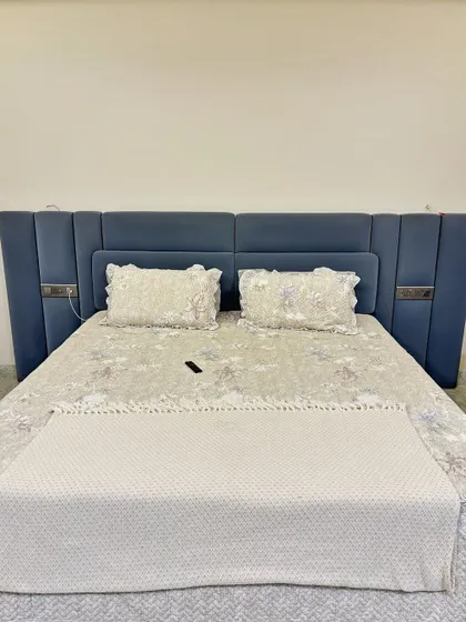 This custom bed features an extra-wide upholstered headboard in a calming blue fabric. The side panels include sleek metallic accents and integrated switches for a clean, modern look.