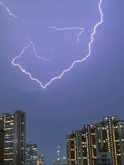 A massive fork of lightning strikes downwards, perfectly framed between the tall buildings of Mumbai at night.