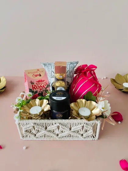 This macrame tray hamper is beautifully styled with golden flower-shaped diyas, a pink potli, and a mix of sweet and savory treats.