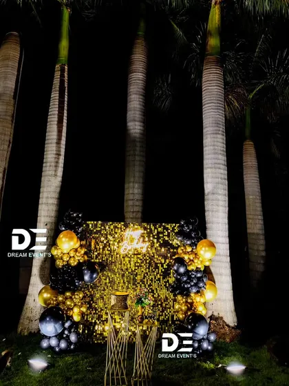 A full view of an outdoor surprise party setup, where a gold shimmer wall and black and gold balloons create a luxurious vibe against the night sky.