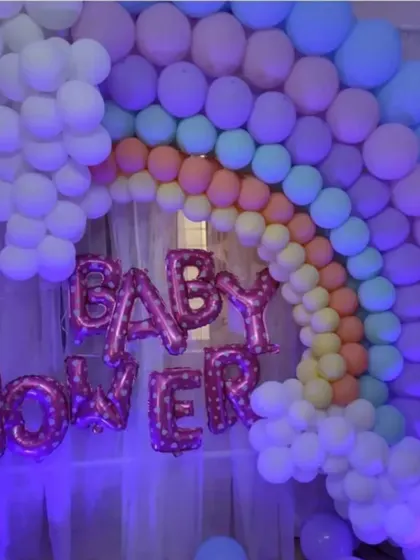 A beautiful rainbow balloon arch against a sheer curtain backdrop for a home baby shower. The 'Baby Shower' foil balloons in pink add a celebratory touch to this colorful and happy setup.