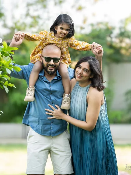 A bright and happy outdoor family portrait. The daughter sits on her father's shoulders, and the whole family radiates joy under the open sky.