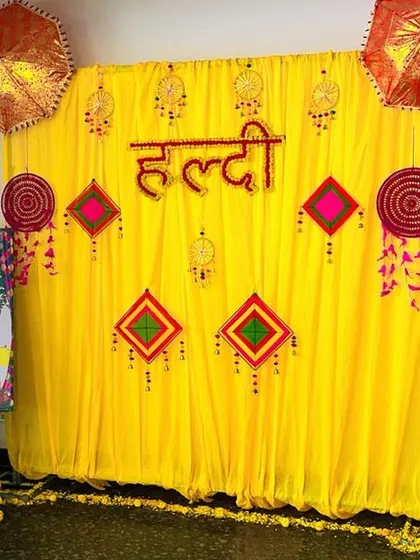 An indoor Haldi and Mehendi corner, personalized with a custom welcome easel board. The backdrop features a traditional yellow curtain with decorative umbrellas and hangings, creating a dedicated space for the ceremonies.