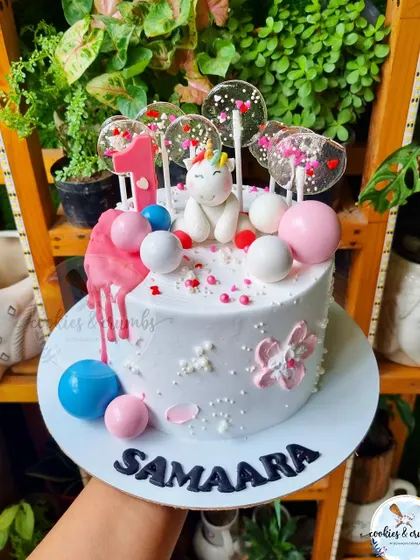A sweet and whimsical unicorn cake for a first month birthday. It's decorated with a fondant unicorn, candy-like lollipops, and a pink drip.