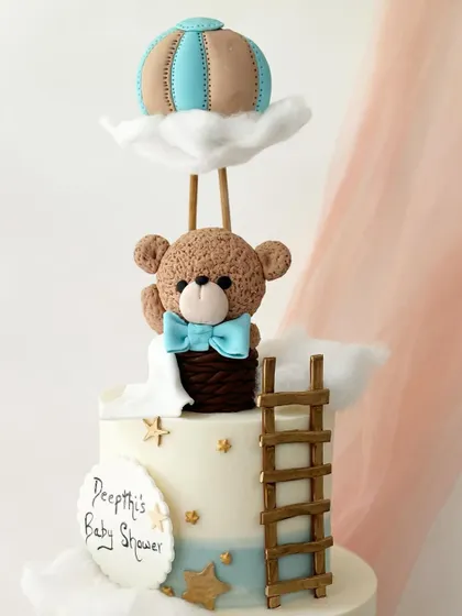 A whimsical "up and away" themed baby shower cake. This design features a teddy bear in a hot air balloon, with fondant clouds and a ladder.