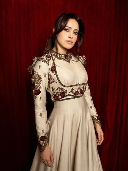 A portrait of Nushrratt Bharuccha with an 'Umrao Jaan' inspired theme. The rich red curtain and traditional outfit are lit to create a regal, cinematic feel.