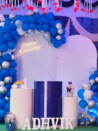 A wider view of the Adhvik's birthday decoration, showing the full arrangement of the balloon arch, backdrop, and props like the decorative bird cage and rainbow sign.