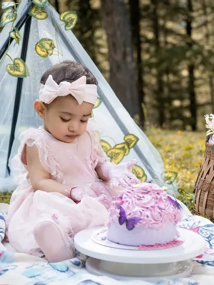 A close-up of the birthday girl intently focused on her cake. The details of the picnic blanket, basket of flowers, and her adorable pink dress add to the charm of this outdoor toddler portrait.