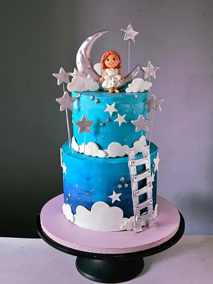 A dreamy, two-tiered cake with a "climb to the moon" theme. It features a blue ombre night sky, edible stars and clouds, a silver ladder, and a charming, hand-sculpted girl figurine sitting on a crescent moon.