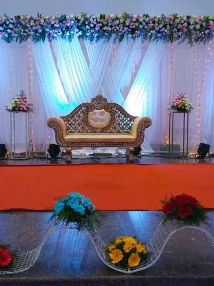 This reception decor features a wide stage with an elegant gold-carved sofa. The backdrop is made of pleated white drapes with blue uplighting, fairy lights, and a full floral border across the top, creating a dreamy and sophisticated look.