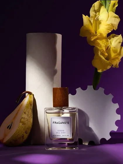 A still life composition for a Fraganote perfume, blending the product with its key ingredients like pear and yellow flowers. The dramatic lighting against a purple backdrop creates a unique, artistic, and memorable product image.