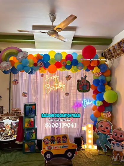 A vibrant Cocomelon theme for a first birthday at home. The setup includes a colorful balloon arch, character cutouts, a light-up number '1', and a toy bus prop.