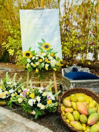 A charming welcome setup for a Haldi ceremony, featuring a custom sign, sunflower arrangements, and a basket of fresh mangoes to tie into the theme.