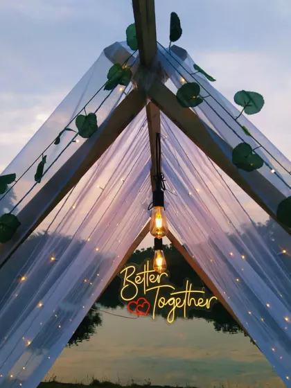 A romantic and intimate anniversary celebration setup in a tent by a lake in Karjat. The warm glow of the 'Better Together' neon sign and fairy lights creates a cozy, unforgettable atmosphere at dusk.