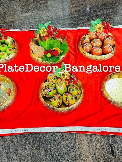 A set of six golden bowls on a red cloth, each filled with different decorated fruits, sweets, and auspicious items for a ceremony.