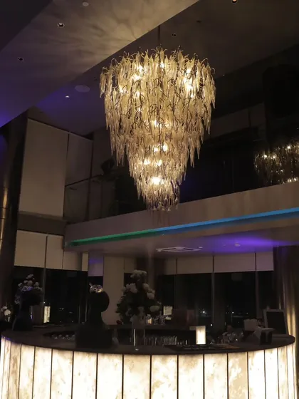 A modern, illuminated bar counter topped by a magnificent, multi-tiered glass leaf chandelier. This combination of custom furniture and statement lighting creates a high-impact bar area for any corporate party.