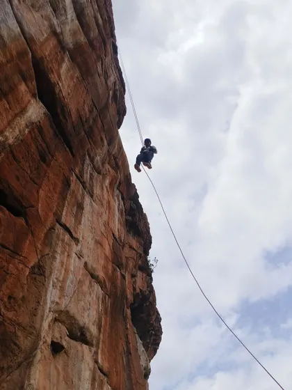 A view from below shows a climber rappelling down a sheer cliff face against the sky.