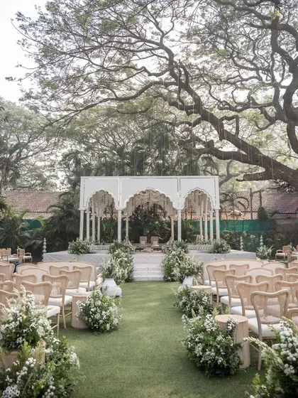 The main wedding mandap, a grand white structure with multiple arches, set under a majestic old tree, blending architectural design with nature.